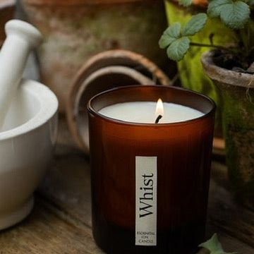Candle with 'Whist' branding on a wooden surface with plants in the background