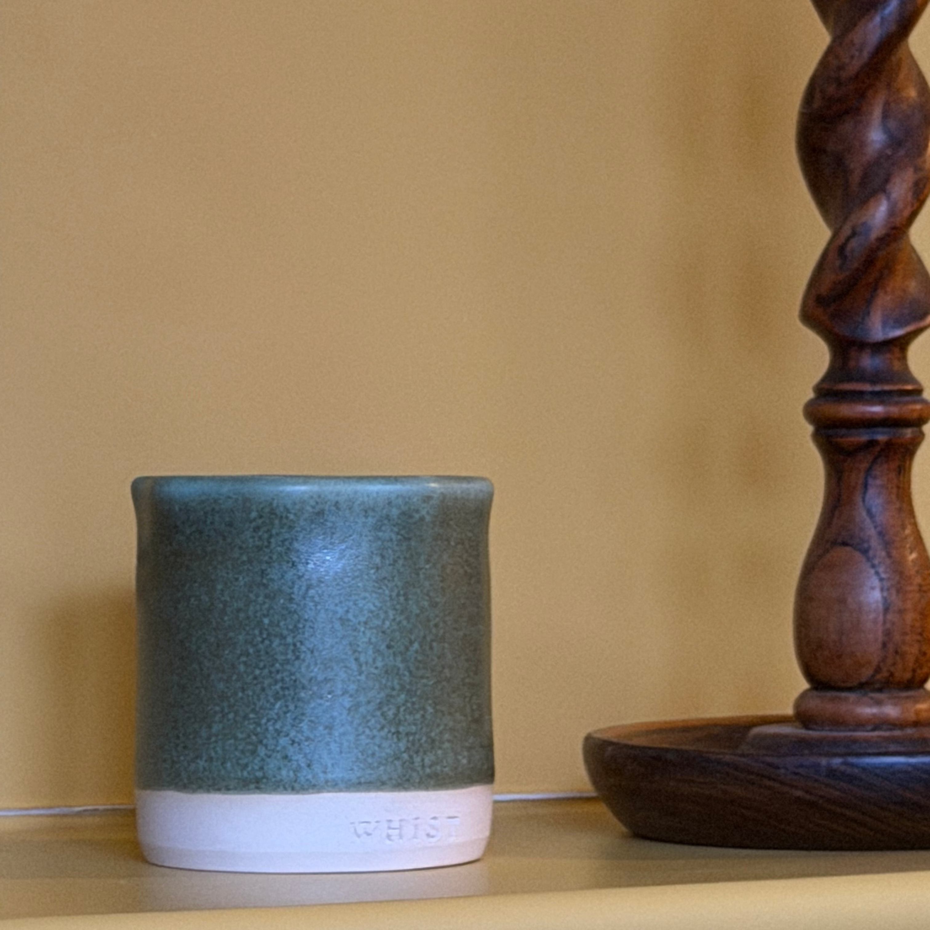 A ceramic candle with a speckled teal-green upper body and a white base embossed with the brand name 'WHIST', resting on a light-colored wooden shelf next to a dark wood spindle.